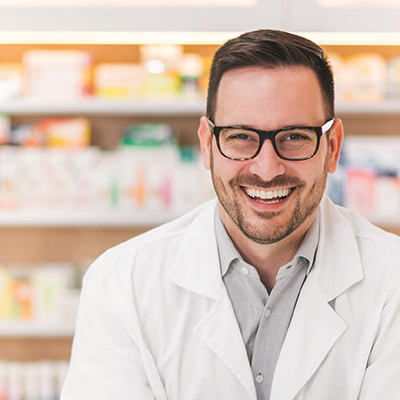Pharmacist with glasses smiling while standing in front of shelves of medication