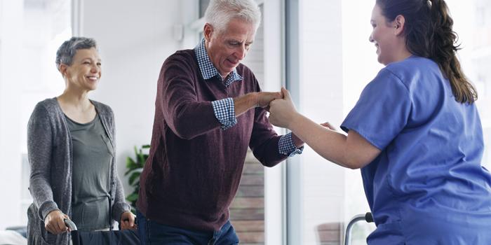 nurse helping an elderly man up to standing position. Woman smiles in the background.