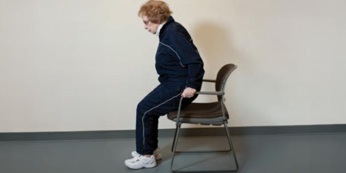 A person is shown gripping the arms of a chair and using their arms to lift bottom off the chair. Holding for one second and lowering back down to perform arm chair pushups.