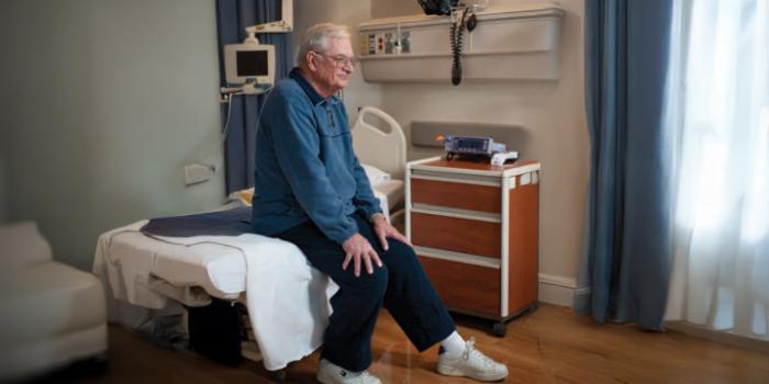 A person sits at edge of bed with feet on floor. Hands are placed on knees to push upward while leaning forward to stand up.